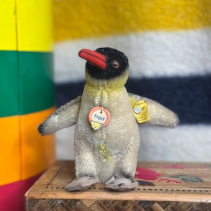 May include: A vintage plush penguin toy with a black head, yellow body, and red beak. The penguin has small, outstretched wings and is standing on a wooden box. It has a tag that says "Peggy".