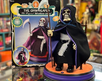 May include: A vintage animated Grim Reaper Halloween decoration. The figure is dressed in a black robe with a purple lining and holds a scythe. The figure is sitting on a rotating base with a purple and orange platform. The box shows the Grim Reaper with the text "The Grim Reaper The Original Motion-Eyes".