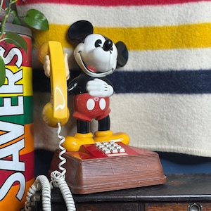 May include: A vintage Mickey Mouse telephone. The phone features a smiling Mickey Mouse figure holding a yellow receiver. The base is brown with a red keypad. The phone is on a dark wooden surface.