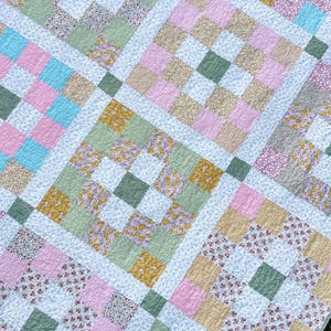 Pode incluir: Uma colcha de retalhos com uma grade de quadrados em vários padrões e cores, incluindo rosa claro, verde sálvia, amarelo mostarda, azul celeste e estampas florais brancas. Os quadrados de tecido são costurados com faixas brancas.