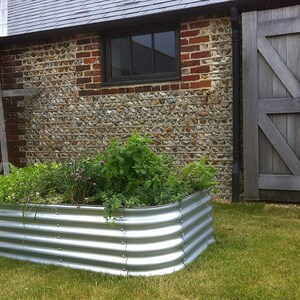 May include: A raised garden bed made of corrugated metal, with a variety of plants growing inside. The bed is positioned in front of a brick and stone wall, with a wooden door to the right.