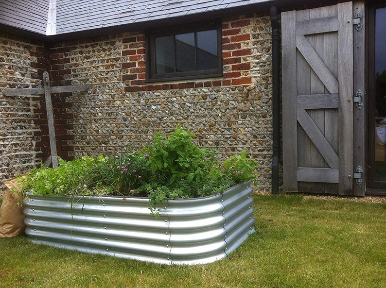 May include: A raised garden bed made of corrugated metal, with a variety of plants growing inside. The bed is positioned in front of a brick and stone wall, with a wooden door to the right.