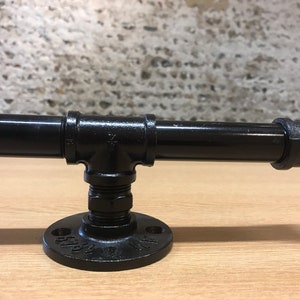 May include: Black metal pipe fitting with a flange and a threaded pipe. The pipe fitting is mounted on a wooden surface.
