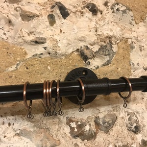 May include: Black metal curtain rod with copper rings. The rod is mounted to a wall with a black metal flange. The flange has the text "1/2" stamped on it.