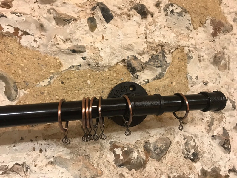 May include: Black metal curtain rod with copper rings. The rod is mounted to a wall with a black metal flange. The flange has the text "1/2" stamped on it.