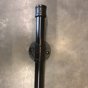 May include: Black metal pipe fitting with a round flange and a threaded end cap. The pipe is attached to a wall.