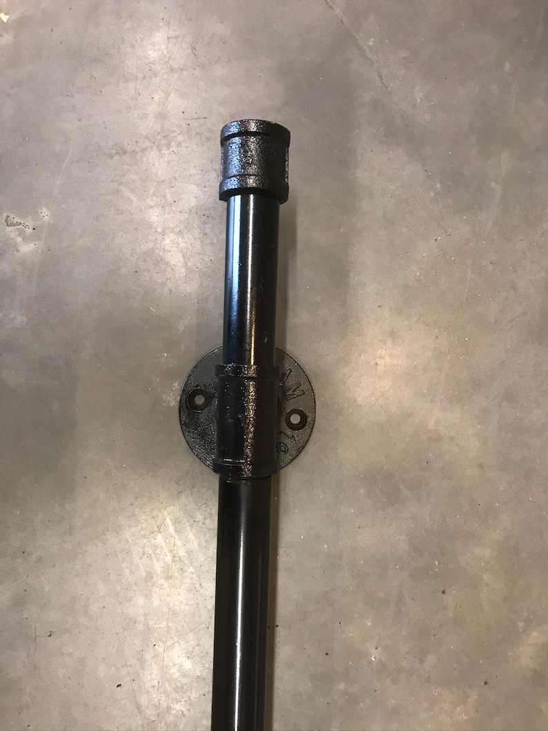 May include: Black metal pipe fitting with a round flange and a threaded end cap. The pipe is attached to a wall.