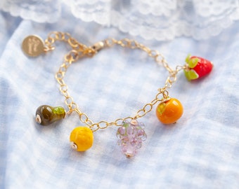 May include: A gold chain bracelet with charms shaped like fruit, including a pear, grapes, a lemon, and a strawberry. The charms are made of glass and have a realistic appearance.