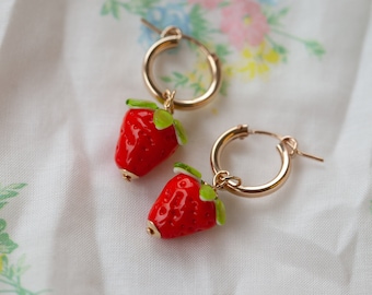 May include: A pair of gold hoop earrings with red and green strawberry charms. The earrings are on a white fabric background with floral patterns.