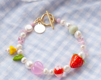 May include: A delicate pearl bracelet with a gold clasp and colorful charms, including a pink heart, a red strawberry, and a green leaf.