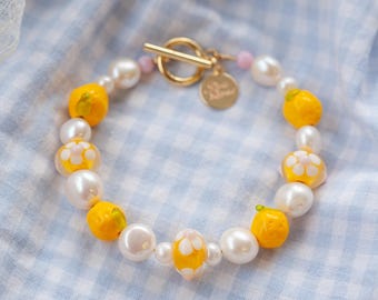 May include: A white pearl and yellow glass bead bracelet with a gold clasp and a small gold charm. The bracelet features yellow glass beads with white flower designs.