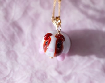 May include: A gold chain necklace with a white and brown glass guinea pig pendant. The pendant has pink accents and black eyes.