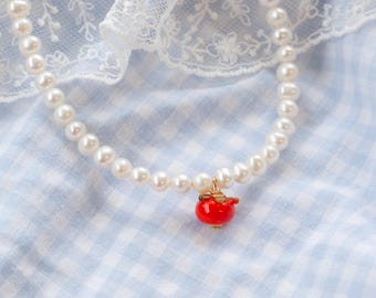 May include: A white pearl necklace with a red glass pendant shaped like a cherry. The necklace is on a blue and white gingham fabric.