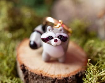 May include: A small glass raccoon charm with a gray and white body, black eyes, and a black and white tail. The charm is sitting on a small wooden slice.