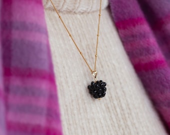 May include: A gold chain necklace with a black blackberry pendant. The necklace is worn over a white sweater and a pink and purple plaid scarf.