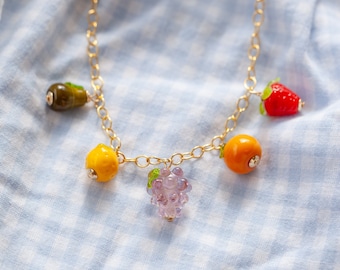 May include: A gold chain necklace with charms of a purple grape cluster, a yellow lemon, a yellow orange, a green pear, and a red strawberry.
