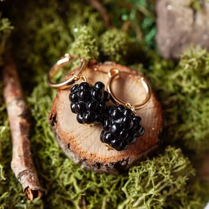 Op de afbeelding: Een paar gouden creolen met zwarte bes-charms. De oorbellen worden getoond op een houten oppervlak bedekt met groen mos.