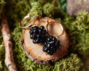 May include: A pair of gold hoop earrings with black berry charms. The earrings are displayed on a wooden surface covered in green moss.