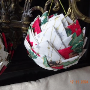 Puede incluir: Dos adornos navideños hechos a mano. Cada adorno es una forma geométrica hecha de tela doblada. La tela tiene un fondo blanco con estrellas doradas y detalles plateados. La tela roja es visible entre los pliegues. También son visibles patrones de árboles de Navidad y poinsettias verdes y rojas.