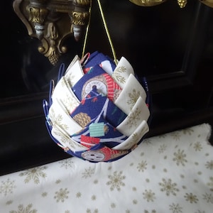 Peut inclure: Une décoration de Noël en tissu bleu et blanc en forme de pomme de pin avec un motif de flocon de neige doré. L'ornement est suspendu à un crochet doré.