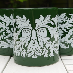 Puede incluir: Tres tazas de cerámica verde con un diseño blanco de la cara de un hombre verde rodeado de hojas.