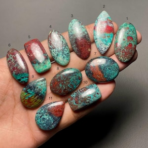 Puede incluir: Una colección de once piedras preciosas pulidas de varias formas, incluyendo ovaladas, en forma de lágrima y rectangulares. Las piedras muestran una mezcla de colores, principalmente verde azulado, rojo y marrón, con patrones y texturas únicos. Las piedras preciosas se sostienen en una mano.