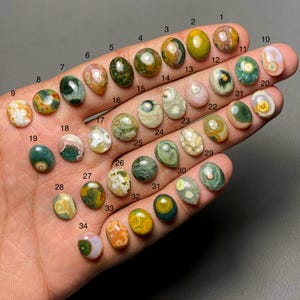 以下が含まれることがあります： 様々な楕円形と丸みを帯びた形の34個の研磨された宝石のコレクション。宝石は、緑、黄、茶色、白など、さまざまな色を示し、ユニークなパターンとテクスチャを持っています。各石には1から34までの番号が付けられています。