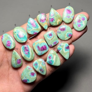 Può includere: Una collezione di sedici pietre preziose lucide di varie forme, tra cui ovali, a goccia e rettangolari. Le pietre presentano un mix di colori, con una base verde chiaro dominante, accenti turchese e inclusioni magenta vibranti. Le pietre preziose sono tenute in una mano.