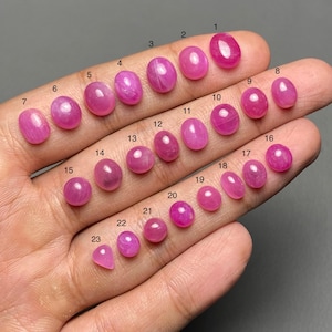May include: A hand displaying 23 polished, oval-shaped, and cabochon-cut gemstones in varying shades of pink and red. Each stone is numbered. The gemstones are likely rubies or similar precious stones.