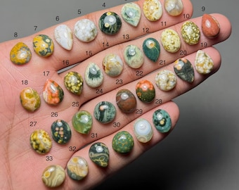 Op de afbeelding: Een verzameling van 33 gepolijste edelsteen cabochons in verschillende vormen en kleuren, waaronder groen, bruin, geel en wit. Elke steen is genummerd en wordt op een hand getoond, wat de verscheidenheid aan natuurlijke patronen en texturen laat zien.