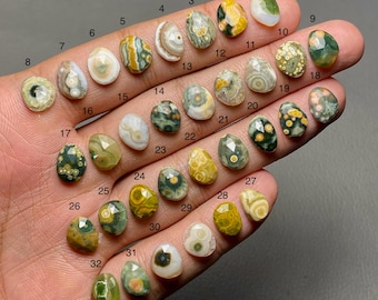 Op de afbeelding: Een verzameling van 32 gepolijste edelsteen cabochons in verschillende kleuren en patronen, waaronder groen, geel, wit en bruin. Elke steen is ovaal van vorm en genummerd. De edelstenen worden op een hand getoond.