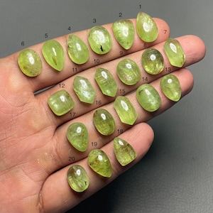 May include: A collection of twenty peridot gemstones in various shapes and sizes, ranging from oval to teardrop. The stones are a translucent, light green color, and are displayed on a hand. Each stone is numbered.