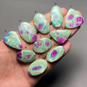 May include: A selection of ten polished gemstones in diverse shapes, such as oval and triangular. The stones display a light green base with striking magenta and teal patterns. The gemstones are held in a hand.