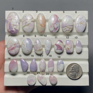 May include: A collection of twenty-two polished gemstones in various shapes and sizes, displayed on a white tray. The stones feature a range of colors, including lavender, pink, and white, with unique veining patterns. A US nickel coin is included for size reference.