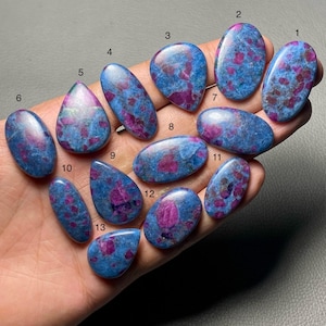 May include: A collection of thirteen polished ruby in zoisite gemstones. The stones vary in shape, including oval, teardrop, and irregular forms. Each stone features a vibrant mix of blue-green zoisite and deep red ruby inclusions.