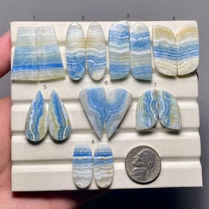 May include: A collection of eight pairs of polished blue aragonite cabochons, each with unique patterns and shapes. The stones display bands of blue, white, and yellow hues. A US nickel is included for size reference.