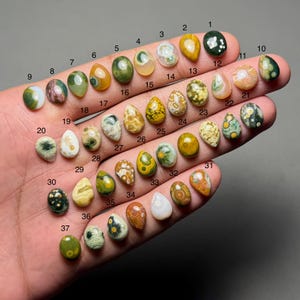 Op de afbeelding: Een verzameling van 37 gepolijste edelstenen in verschillende vormen en kleuren, waaronder groen, geel, bruin en wit. Elke steen is genummerd en wordt in een hand gehouden, waarbij hun unieke patronen en texturen worden getoond. De edelstenen variëren in grootte en vorm.