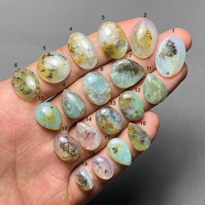 May include: A collection of nineteen polished gemstones in various shapes and colors, including oval, teardrop, and round. The stones feature a mix of translucent and opaque qualities, with colors ranging from pale blue and green to yellow and brown. Some stones have internal patterns.