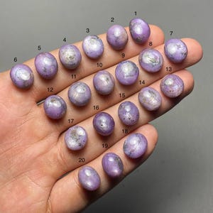May include: Twenty oval-shaped, lavender-coloured gemstones are displayed. Each stone has a smooth, polished surface with subtle variations in colour and some with silver inclusions. The gemstones are numbered from 1 to 20.