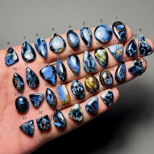 Op de afbeelding: Een verzameling van drieëndertig gepolijste pietersiet edelstenen in verschillende vormen, waaronder druppels, ovalen en rechthoeken. De stenen vertonen een mix van blauwe, gouden en zwarte kleuren, met zichtbare patronen en texturen. De edelstenen worden in een hand gehouden.