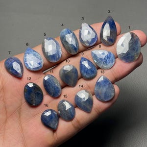 May include: Sixteen blue sapphire gemstones of different shapes, including oval, pear, and teardrop. The faceted gemstones display a range of blue hues and internal clarity. The gems are held in a hand against a dark background.