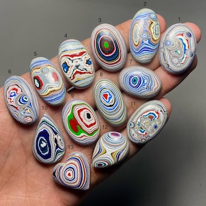 May include: A selection of thirteen polished stones in diverse shapes, including oval, teardrop, and heart. The stones display complex patterns of swirling colours, such as red, blue, green, and white, creating a unique and artistic appearance.