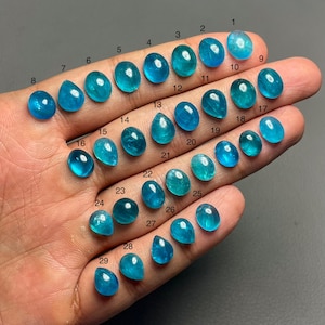 Puede incluir: Una colección de 29 piedras preciosas azules de varias formas, incluyendo ovaladas y en forma de lágrima. Cada gema está numerada del 1 al 29. Las gemas varían en tonos de azul, desde turquesa claro hasta verde azulado intenso. Las piedras están en una mano.