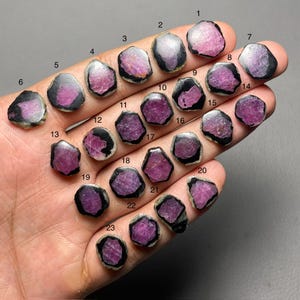 Puede incluir: Una colección de veintitrés piedras preciosas en bruto, cada una con un centro magenta intenso y un borde oscuro e irregular. Las piedras se exhiben sobre una superficie clara, mostrando su estado natural y sin pulir. Las piedras preciosas están numeradas del 1 al 23.