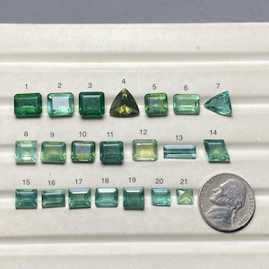 Puede incluir: Una colección de veintiuna piedras preciosas en varias formas y tonos de verde y verde amarillento, dispuestas sobre una superficie clara. Se incluye una moneda de níquel estadounidense para comparar el tamaño. Las piedras preciosas están numeradas del 1 al 21.