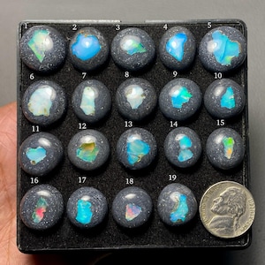 May include: A collection of nineteen dark gray, spherical stones, each with a unique, colorful opal inlay. The opals display a range of colors, including blue, green, and red. A nickel coin is included for size reference.