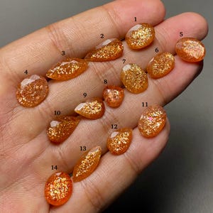 Puede incluir: Una colección de catorce piedras preciosas de piedra solar facetadas en varias formas, incluyendo cortes ovalados, de pera y marquesa. Cada piedra de tono naranja presenta inclusiones metálicas brillantes, etiquetadas con números negros del uno al catorce.