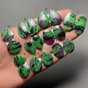 May include: A collection of faceted zoisite gemstones with ruby inclusions, displaying vibrant green, black, and magenta patterns. The stones are arranged in groups labeled with numbers 1 through 9, showcasing various teardrop, oval, and round shapes.