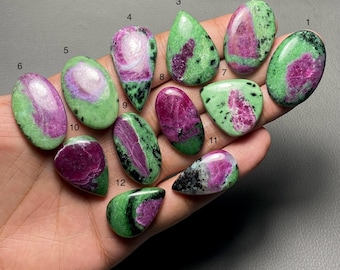 Può includere: Una collezione di dodici pietre preziose lucidate di rubino in zoisite di varie forme. Le pietre presentano un mix di colori verdi, rosa e neri. Le pietre preziose sono tenute in una mano, mostrando i loro motivi e le loro trame uniche.