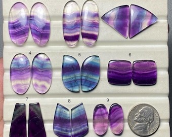 Puede incluir: Una colección de nueve pares de piedras preciosas de fluorita pulidas en varias formas. Las piedras muestran bandas de tonos morados, azules y claros. Las formas incluyen óvalos, triángulos y cuadrados. Se incluye una moneda de níquel estadounidense como referencia de tamaño.